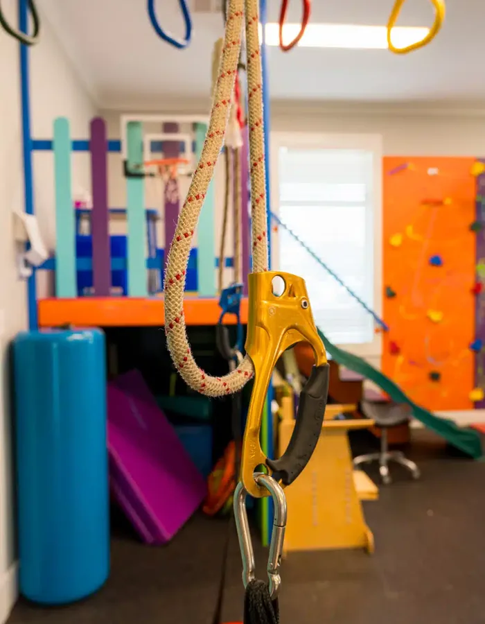 Climbing rope and carabiner hanging in the pediatric sensory gym with padded mats and a climbing wall behind